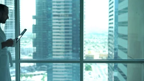 Man in bathrobe reading news on tablet computer and drinking coffee by window at Stock Footage 70708125