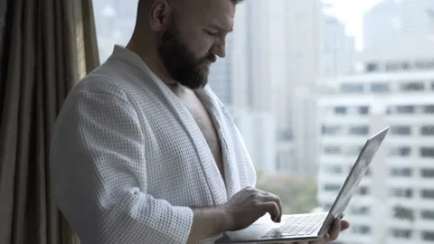 Man in bathrobe standing next to the window and using notebook Stock Footage 88158831