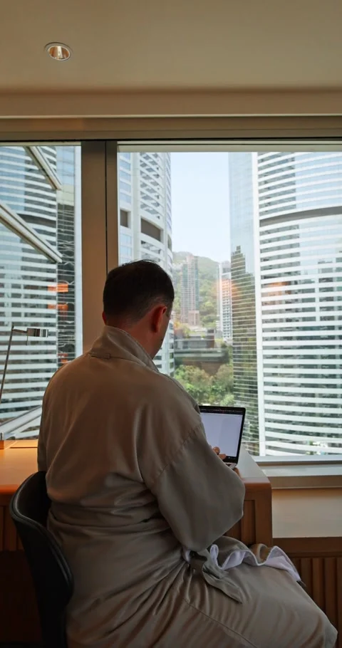 Man in bathrobe working on laptop by window with urban skyline, repres Stock Footage 326948157