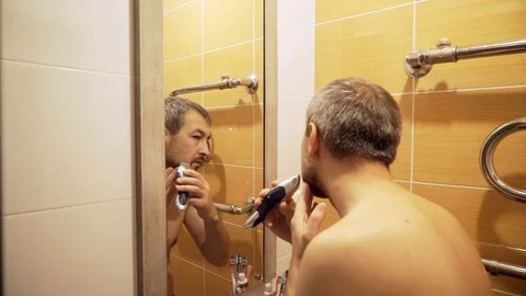 The man in the bathroom in front of the mirror cuts his beard with a trimmer. Stock Footage 82227123