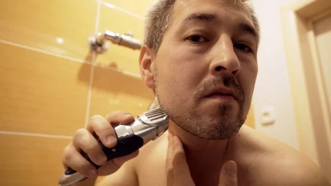 The man in the bathroom in front of the mirror cuts his beard with a trimmer. Stock Footage 82568492