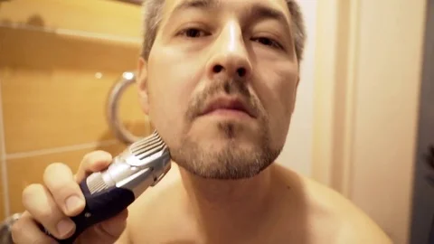 The man in the bathroom in front of the mirror cuts his beard with a trimmer. Stock Footage 82575775