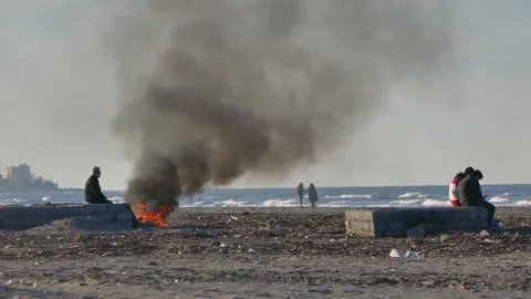 Man by beach fire as thick black smoke rises over polluted shoreline Stock Footage 320744800