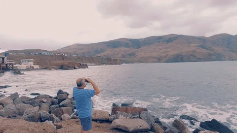 A man on the beach looking through binoculars on the mountains. Waves hit the Stock Footage 97863188