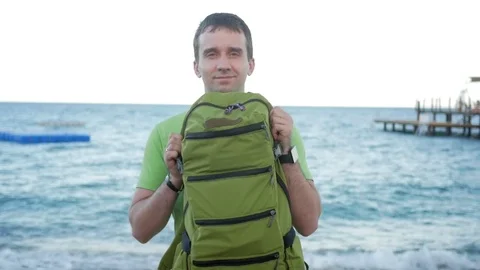 Man on the beach shows a green backpack and smiles at the camera. He turns and Stock Footage 76471793