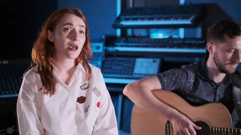 Man with a beard accompanies a red haired girl on a guitar in a music studio Stock Footage 138264874