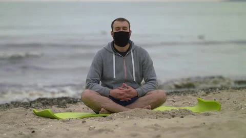 Man with Beard and Face Mask Meditating on Seashore at Dusk - Wide Shot Stock Footage 130731193