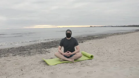 Man with Beard and Face Mask Meditating on Seashore at Dusk - Wide Dolly Shot Stock Footage 130734411