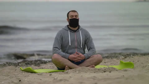 Man with Beard and Face Mask Meditating on Seashore at Dusk - Wide Rack Focus Stock Footage 130738529