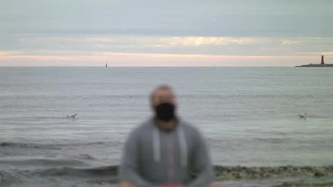 Man with Beard and Face Mask Meditating on Seashore at Dusk - Wide Rack Focus Stock Footage 130738845