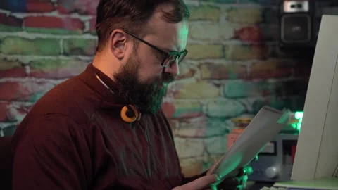Man With Beard and Glasses Looking at Papers in Front of Desktop Computer From Stock Footage 305833813