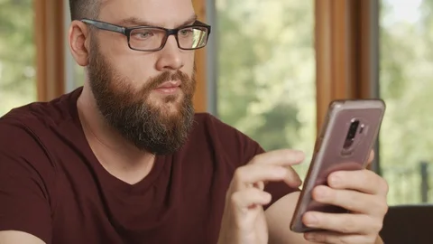 Man with beard and glasses scrolls and reads cell smartphone at home 動画素材 127935000