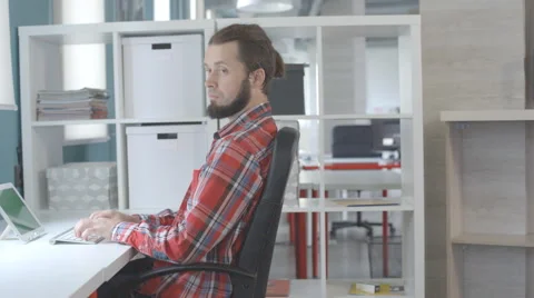 A man with a beard and top knot sits close to chair in office and typing on Stock Footage 67167830