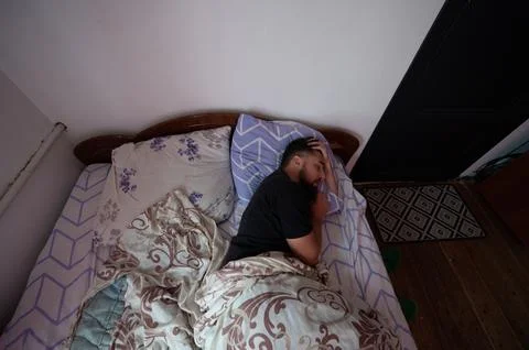 A man with a beard is asleep in a bed with patterned sheets and pillows. His  Stock Photos