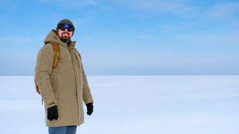 Man with beard in beanie, ski goggles and winter jacket smiles at camera whil 스톡 사진