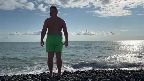 Man with beard, big belly, green shorts on sea pebble beach looks around Stock Footage 332561051