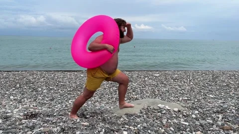 Man with beard, big belly in yellow shorts with magenta inflatable ring beach Stock Footage 304717166