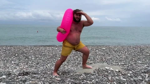 Man with beard, big belly, yellow shorts, swim pink ring on sea pebble shore Stock Footage 332573513