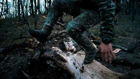 A man with a beard in camouflage with a weapon jumps over a large fallen tree Stock Footage 128121889