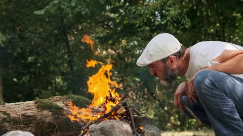 A man with a beard in a cap makes a fire in the woods Stock Footage 296043920