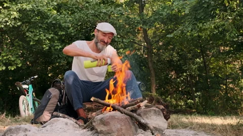 A man with a beard in a cap outdoors by the fire pours tea and coffee from a Stock Footage 148337874