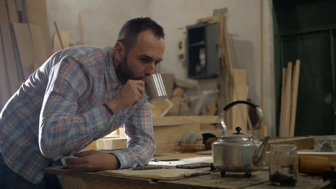 A man with a beard in a carpentry workshop drinking tea and doing calculation Stock Footage 99592082