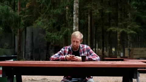 Man with beard checks phone while sitting at wooden table in forest setting Stock Footage 322124193