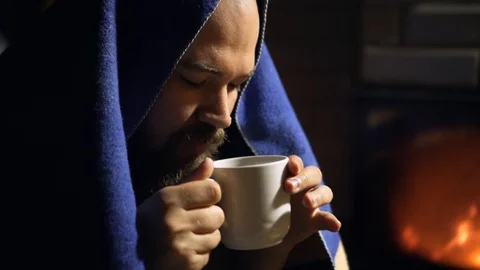 Man with beard covered with blanket drinks hot tea from a cup near fireplace Stock Footage 125093143