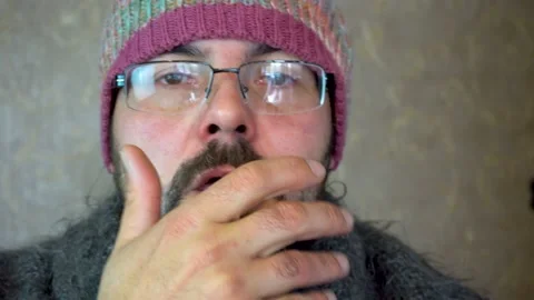 A man with a beard in glasses and a colored hat coughs. 4K Stock Footage 118677119