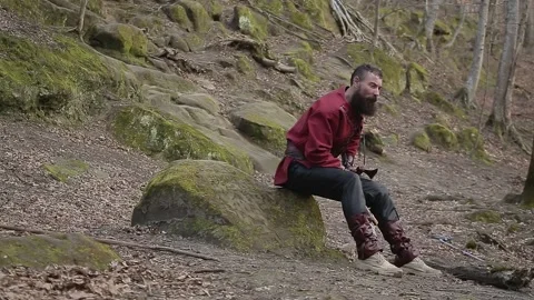 Man with beard in the image of Viking with axe sits on stone Video stock 132992196