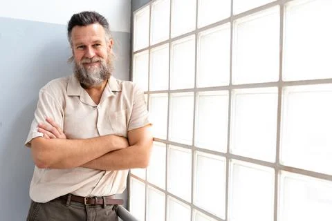 Man with beard looking at camera with arms crossed next to window. Copy space. Stock Photos