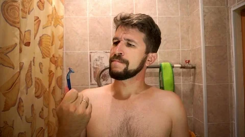 A man with a beard looking at a disposable razor dissatisfied. 库存影片 93819451
