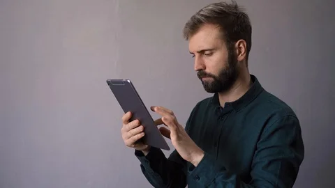A man with a beard looks something in his phone against the background of a whit Stock Footage 128360012