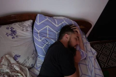 A man with a beard is lying in bed with patterned pillows and bedding, eyes c Stock Photos