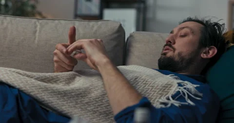 Man with beard is lying on sofa and covered with blanket. The brunette holds Stock Footage 218324471