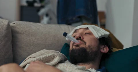 Man with beard is lying on the sofa and taking temperature. He holds thermometer Vídeo Stock 218332718