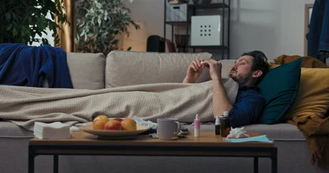Man with beard is lying on sofa covered with blanket and taking temperature. He Stock Footage 218318081