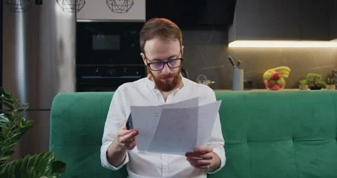 Man beard managing finances, using calculator sitting on sofa in kitchen Stock Footage 241833055