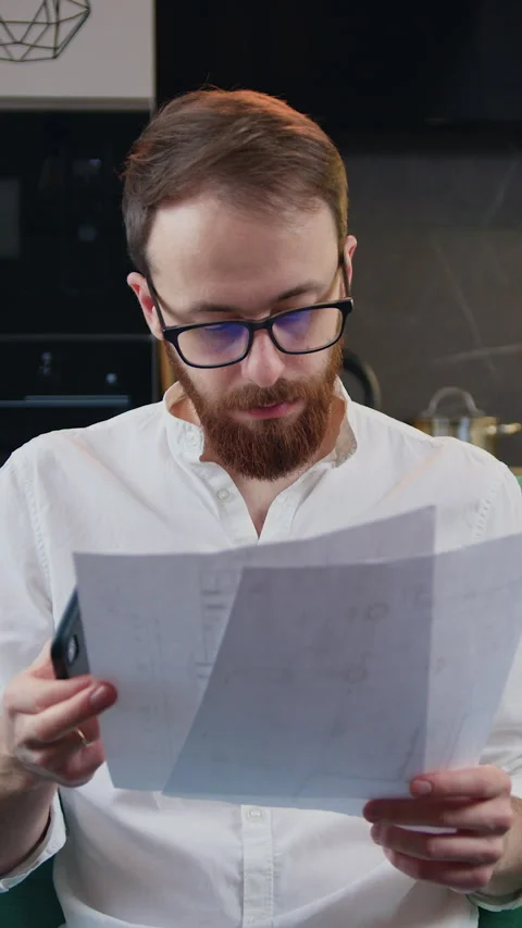 Man beard managing finances, using calculator sitting on sofa in kitchen. Financ Stock Footage 333196423