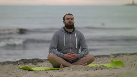 Man with Beard Meditating on Seashore at Dusk - Wide Rack Focus Shot Stock Footage 130732880