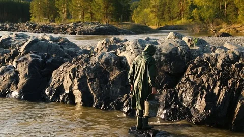 Man beard mustache raincoat is standing near a river with a bucket Stock Footage 111499850