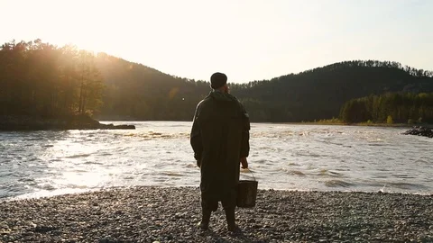 Man beard mustache raincoat is standing near a river with a bucket Stock Footage 111499869