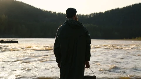 Man beard mustache raincoat is standing near a river with a bucket Stock Footage 111499873