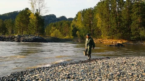 Man beard mustache raincoat is walks near a river with a bucket Stock Footage 111499889