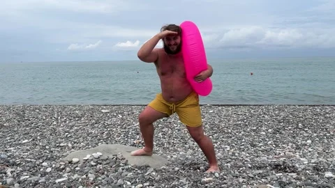 Man with beard, pink inflatable ring in yellow shorts on pebble beach Stock Footage 308128012
