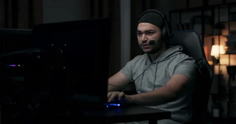 Man with beard plays online game on computer. Guy is wearing cap and headphones Stock Footage 214644277