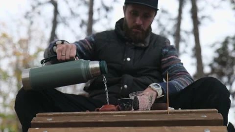 A man with a beard pours boiling water from a thermos into a clay teapot Stock Footage 229860800