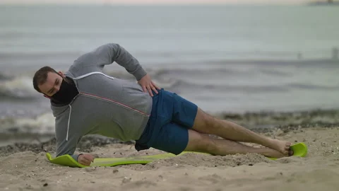 Man with Beard in Protective Face Mask Performing Side Elbow Plank on Seashore Stock Footage 130734558
