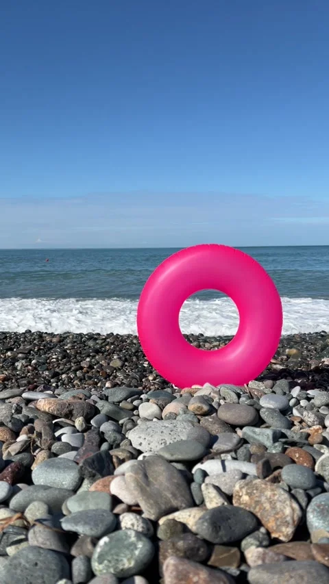 Man with beard runs along pebble beach and catches pink swim ring Video stock 312629079