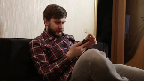 Man with a beard sits on the couch and looks into a smartphone Stock Footage 86159809